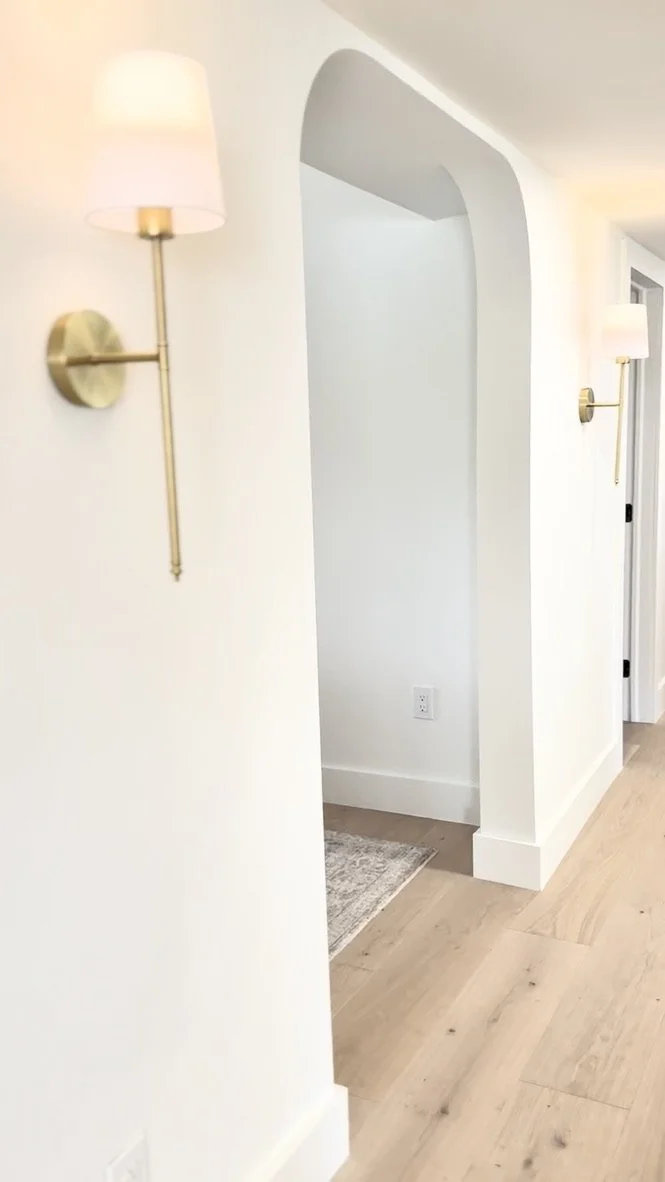 View of a hallway with wall-mounted lamps on each side, white walls, light wood flooring, and an arched doorway leading to another room.