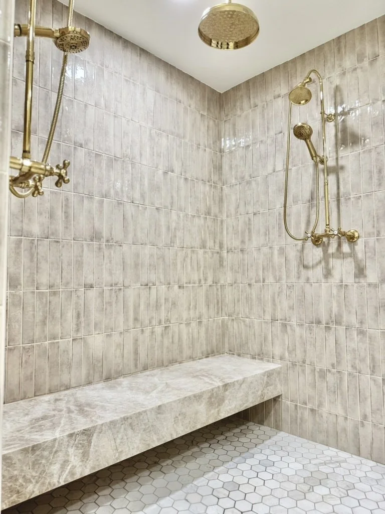 A walk-in shower with beige tiled walls, a built-in marble bench, and two gold-colored showerheads mounted on the wall.
