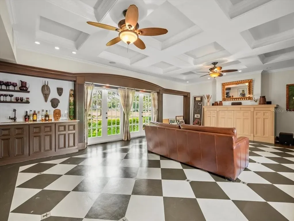 A spacious living room with a checkered black and white tile floor, a large window with curtains, a leather sofa, ceiling fans, and built-in cabinets along the walls.