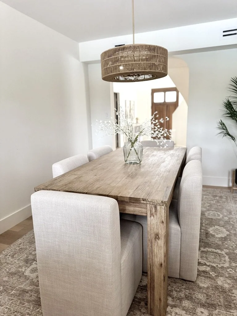 A dining room with a wooden table, beige upholstered chairs, a glass vase with white flowers, a woven rattan pendant light, and a patterned area rug.