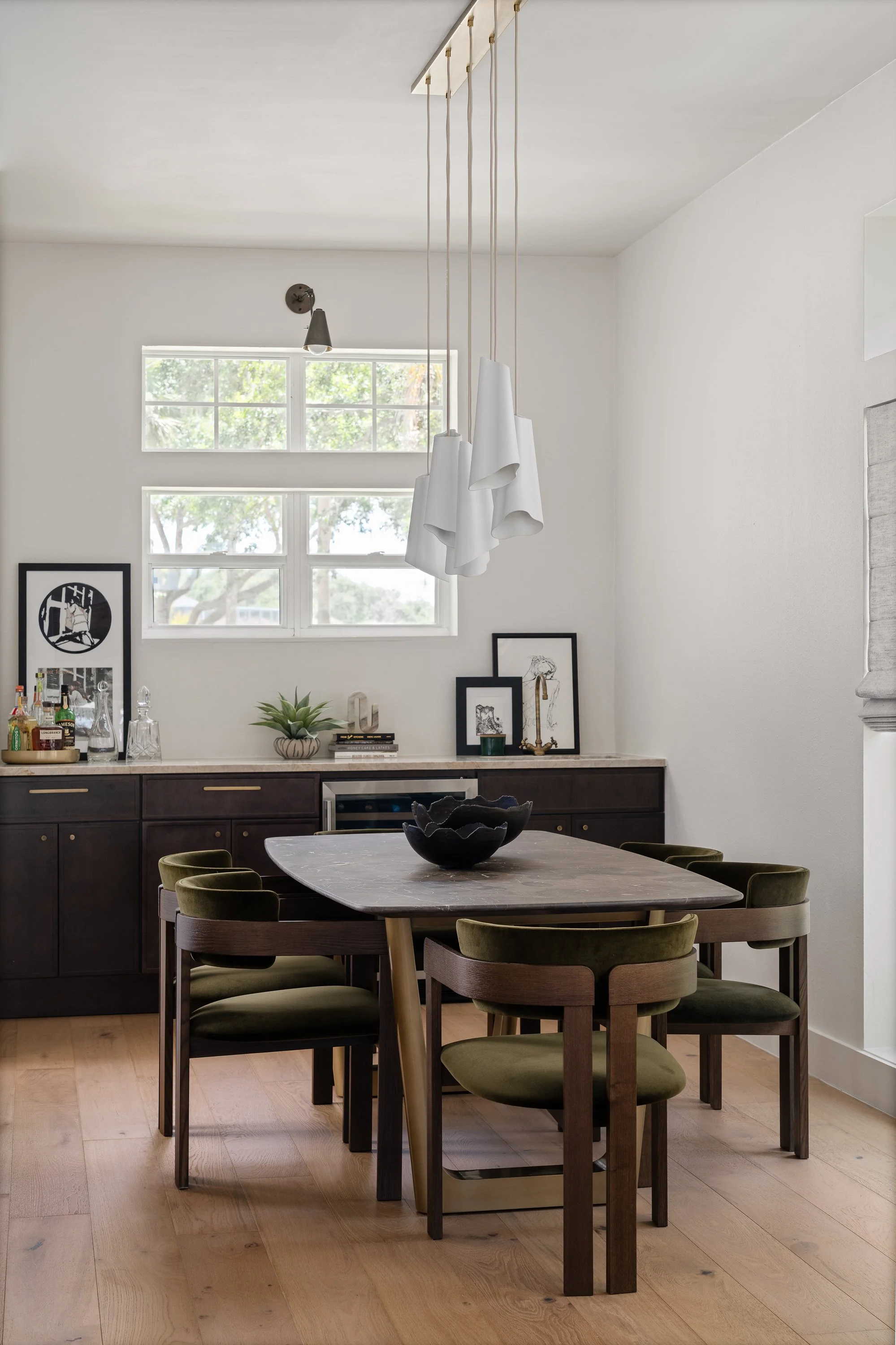 Modern dining room with dark wood table, six green upholstered chairs, a black decorative bowl, and a sideboard with framed artwork, plant, and alcohol bottles. Bright window on white wall, contemporary hanging light fixture, and light hardwood floor