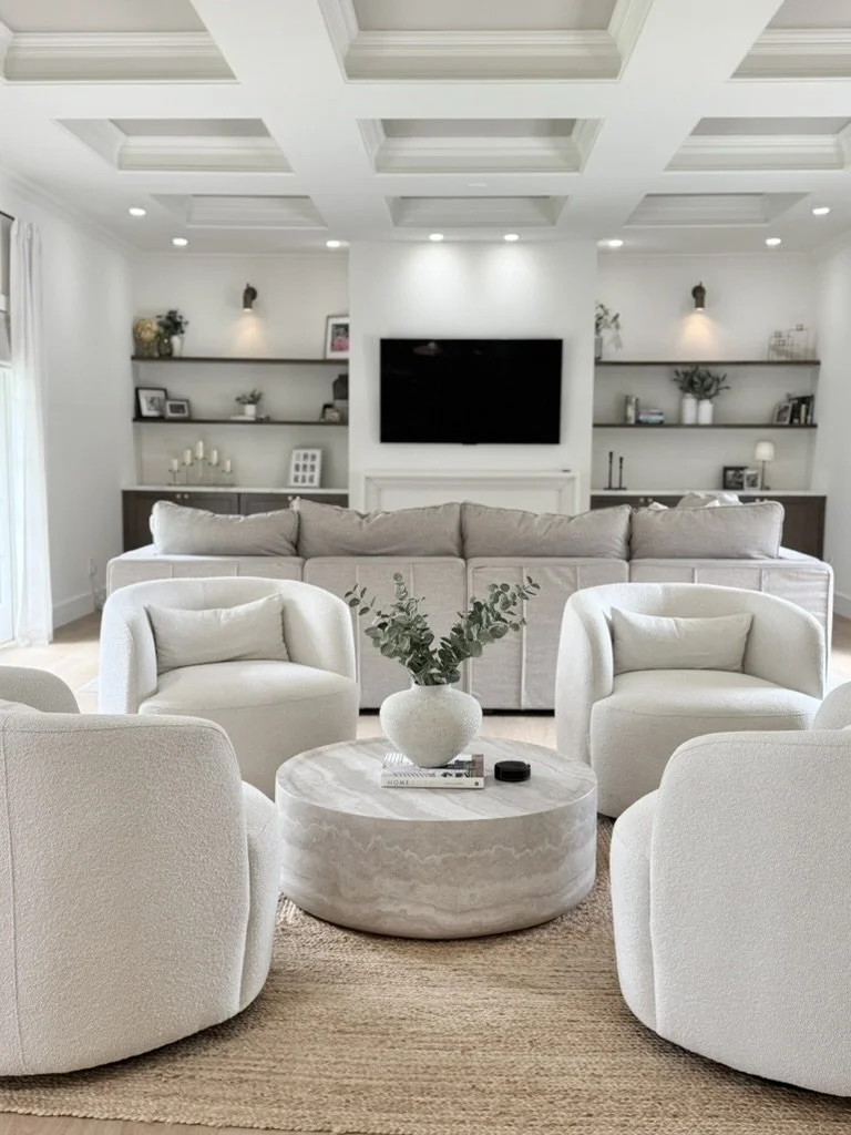Living room with white chairs, a gray couch, a marble coffee table with a vase of greenery, and a wall-mounted TV, with built-in shelves on either side.