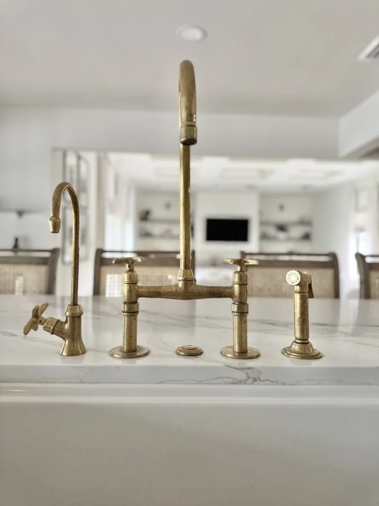 Close-up of three brass kitchen faucets and a sink stopper on marble countertop in a modern kitchen.