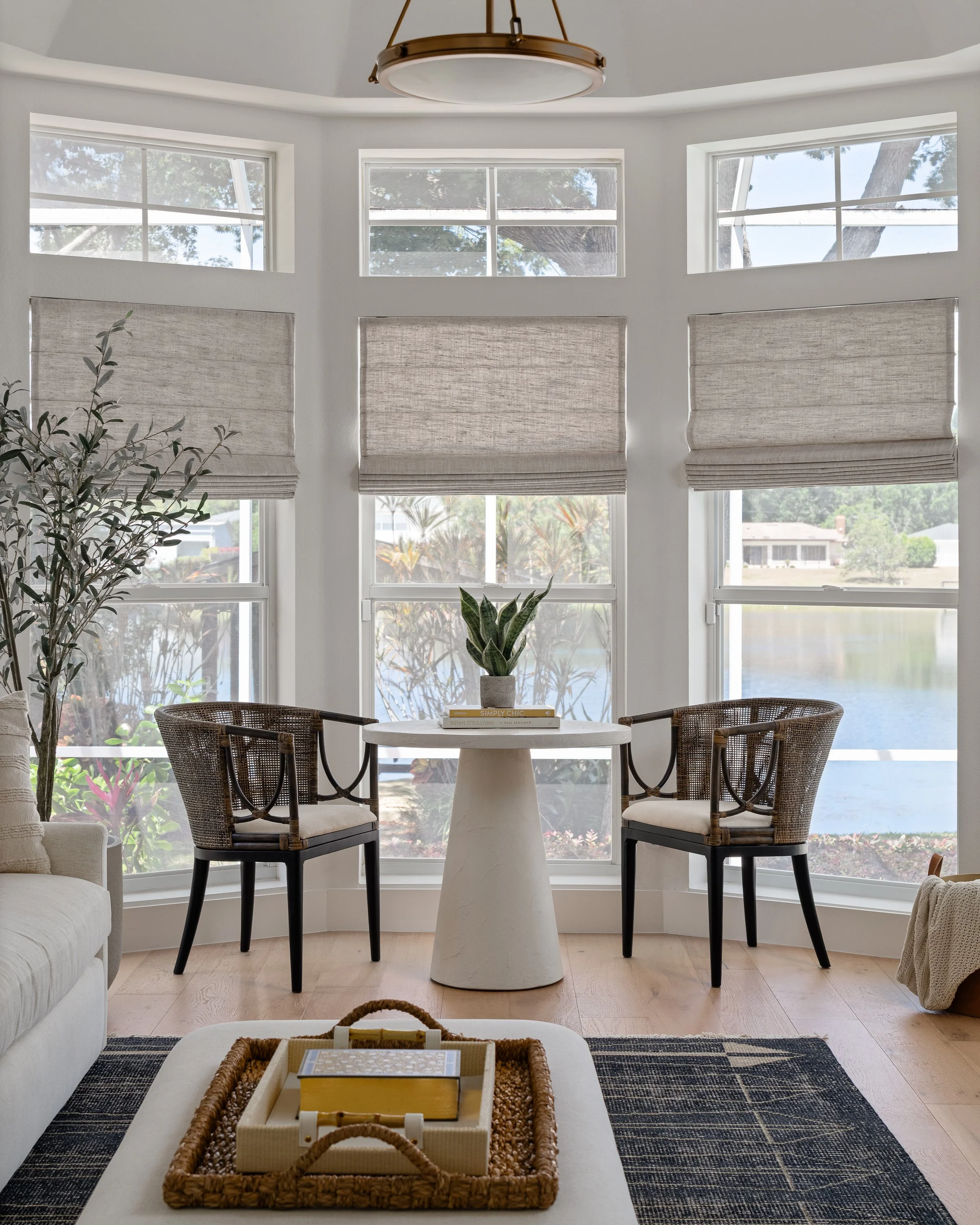 A cozy living room with a bay window and beige Roman shades, a round table with a potted plant and books, two wicker chairs, a sofa, and a coffee table with a tray holding decorative objects.
