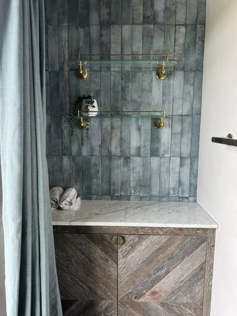 Bathroom with blue tiled wall, two glass shelves with brass supports, a white vase with green plants, and folded towels on marble countertop with wooden cabinets.