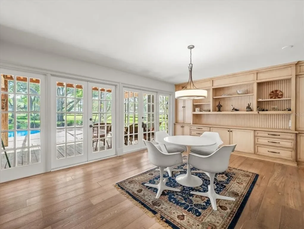 Dining room with French doors leading to a deck and backyard, featuring a white round table with four matching chairs, a decorative rug, and a built-in wooden wall unit with shelves and cabinets.