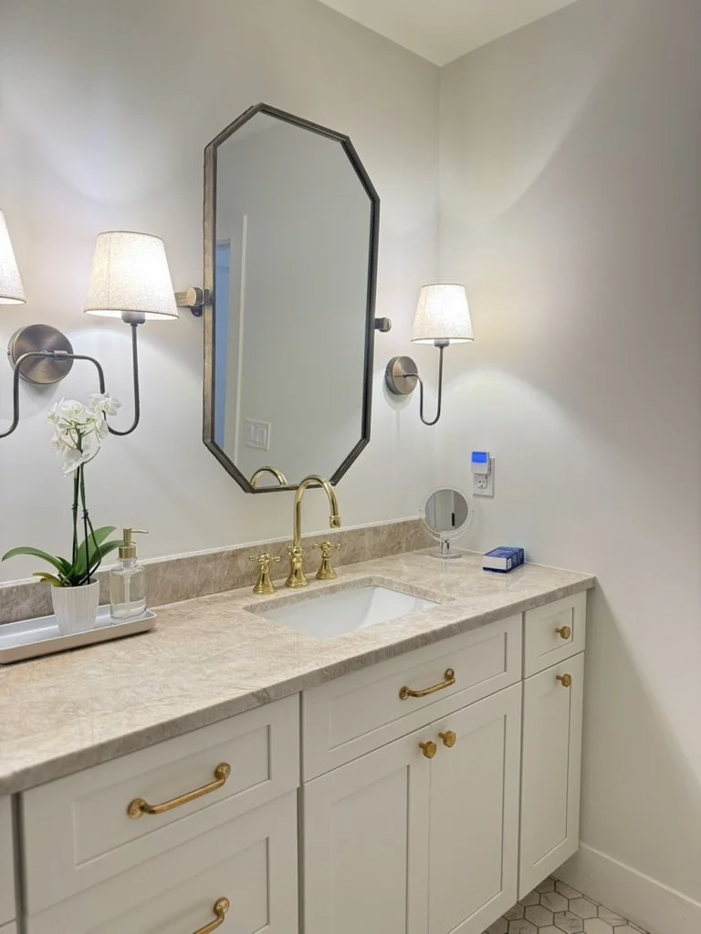 Bathroom vanity with beige marble countertop, white cabinets with gold handles, a gold faucet, a small mirror, wall-mounted lamps with white shades, a potted orchid, soap dispenser, and a box of tissues.