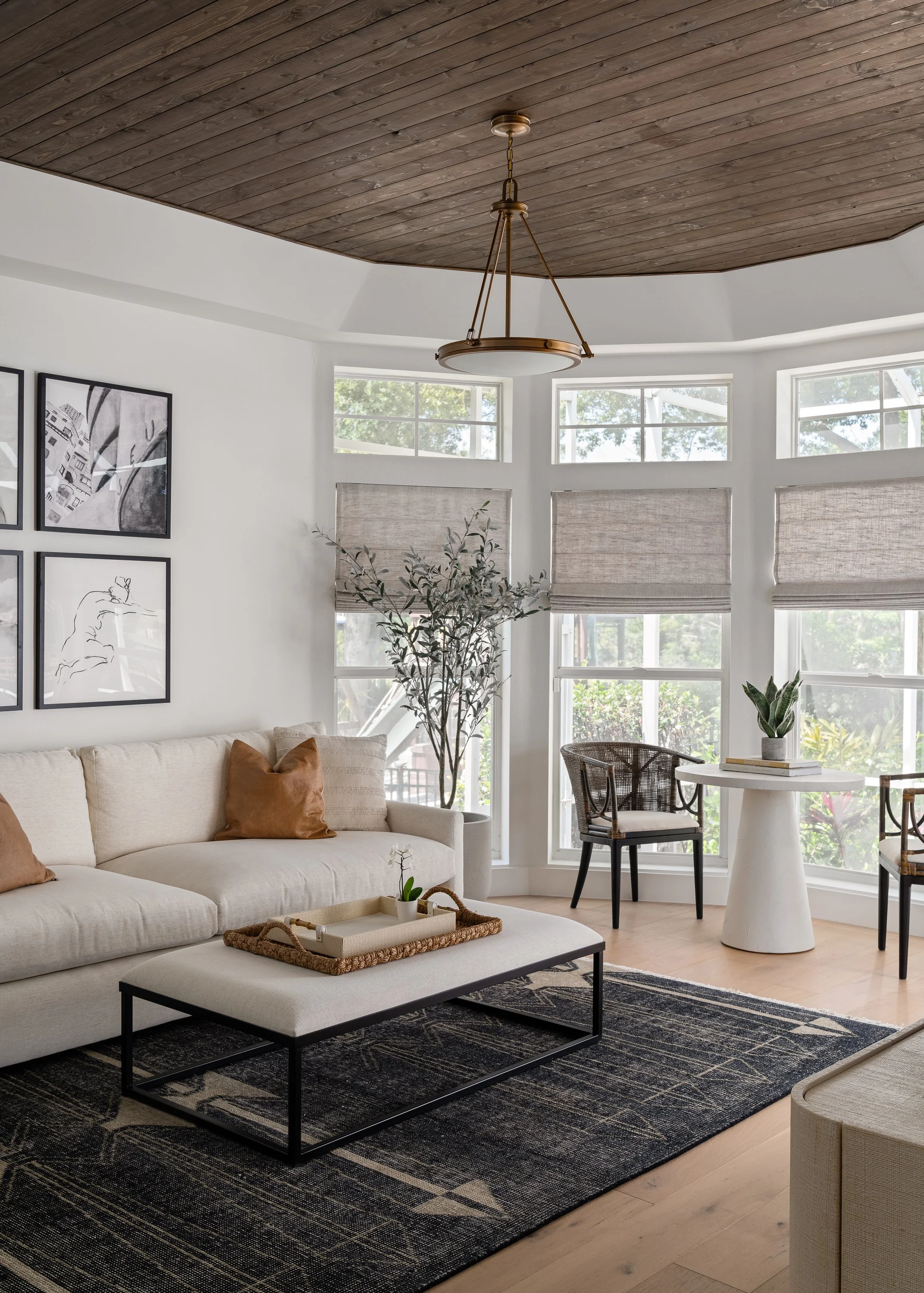 Bright living room with a beige sofa, black framed artwork, large bay windows with blinds, a black and beige rug, and a wooden ceiling with a brass chandelier.
