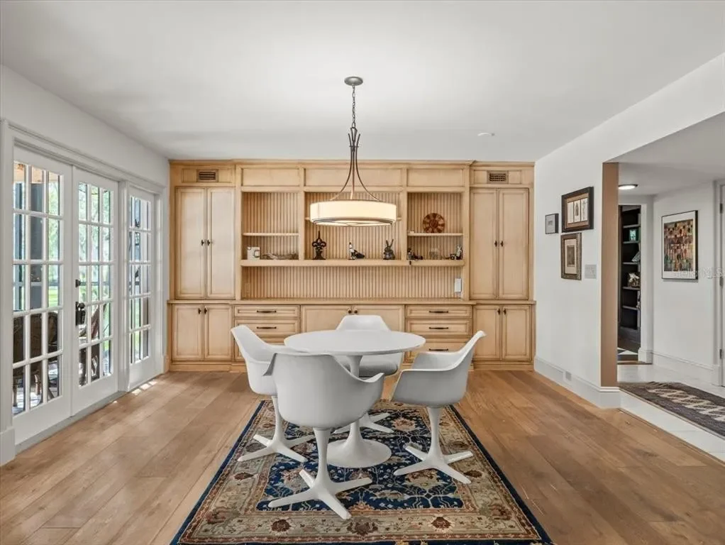 Dining room with round white table, five white chairs, built-in wooden shelves and cabinets, chandelier, sliding glass doors, hardwood floors, and framed artwork on the walls.