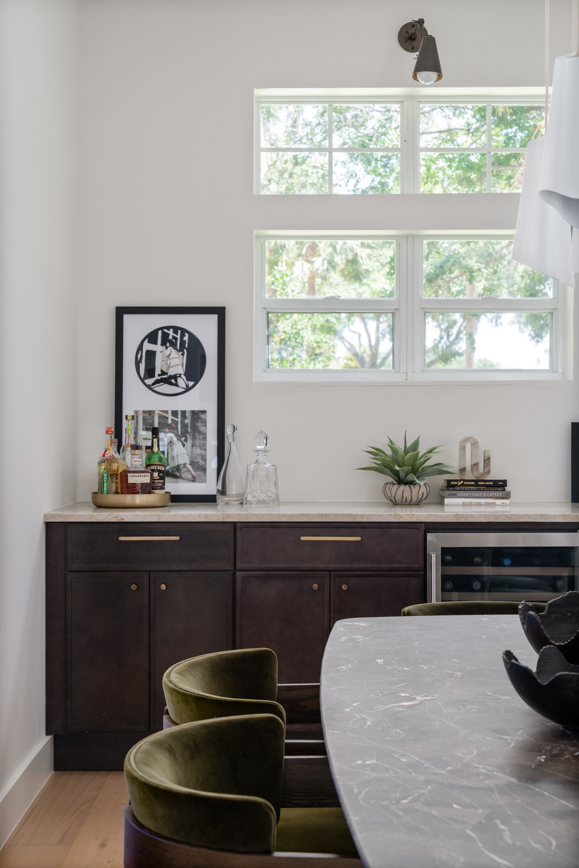 Modern kitchen or dining area with large window, dark wood cabinetry, marble countertop, and green velvet chairs around a marble table. Decor includes books, artwork, a tray with bottles, and a plant.