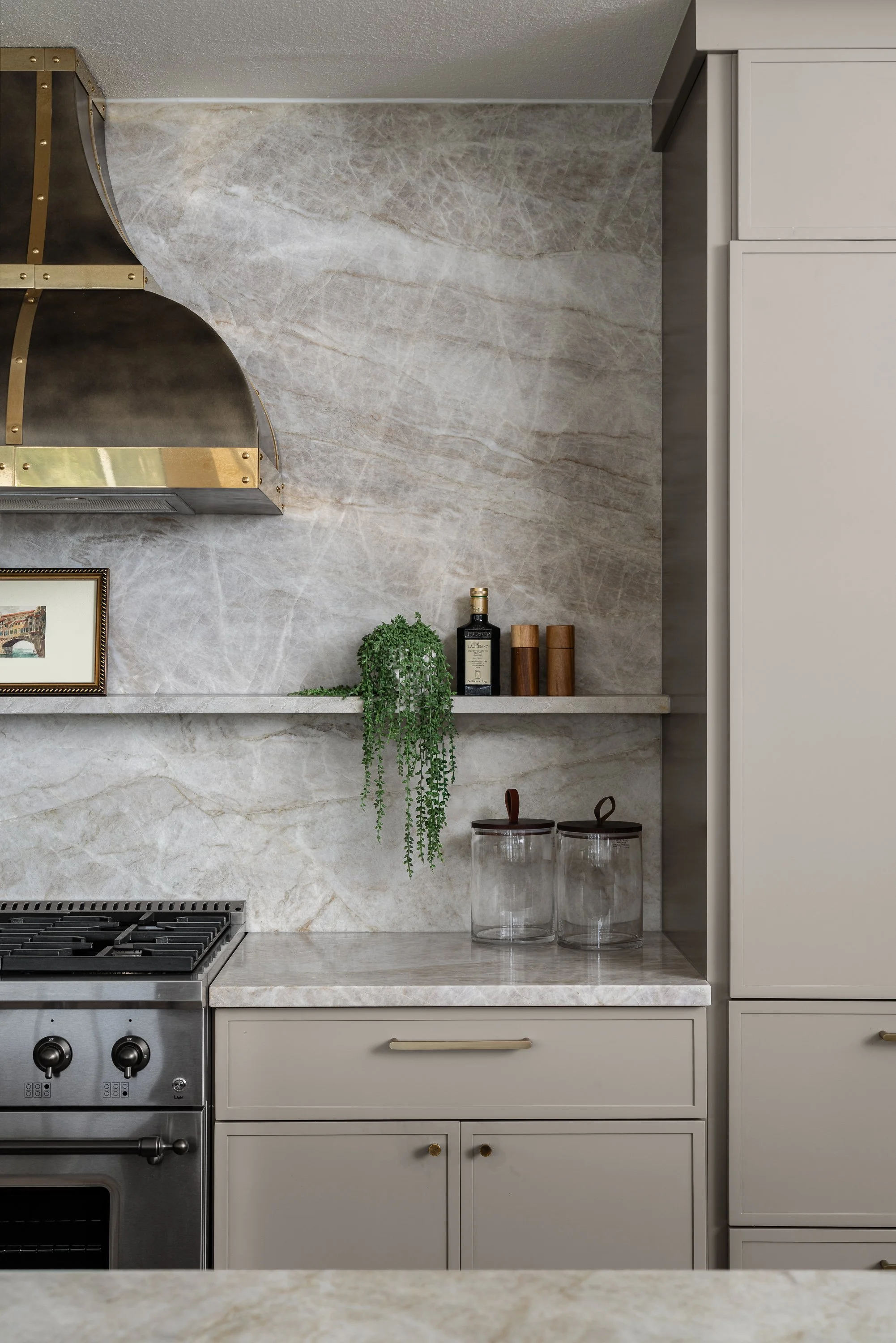 Modern kitchen with marble backsplash, beige cabinets, a stainless steel stove, glass jars on the counter, and a small framed picture.