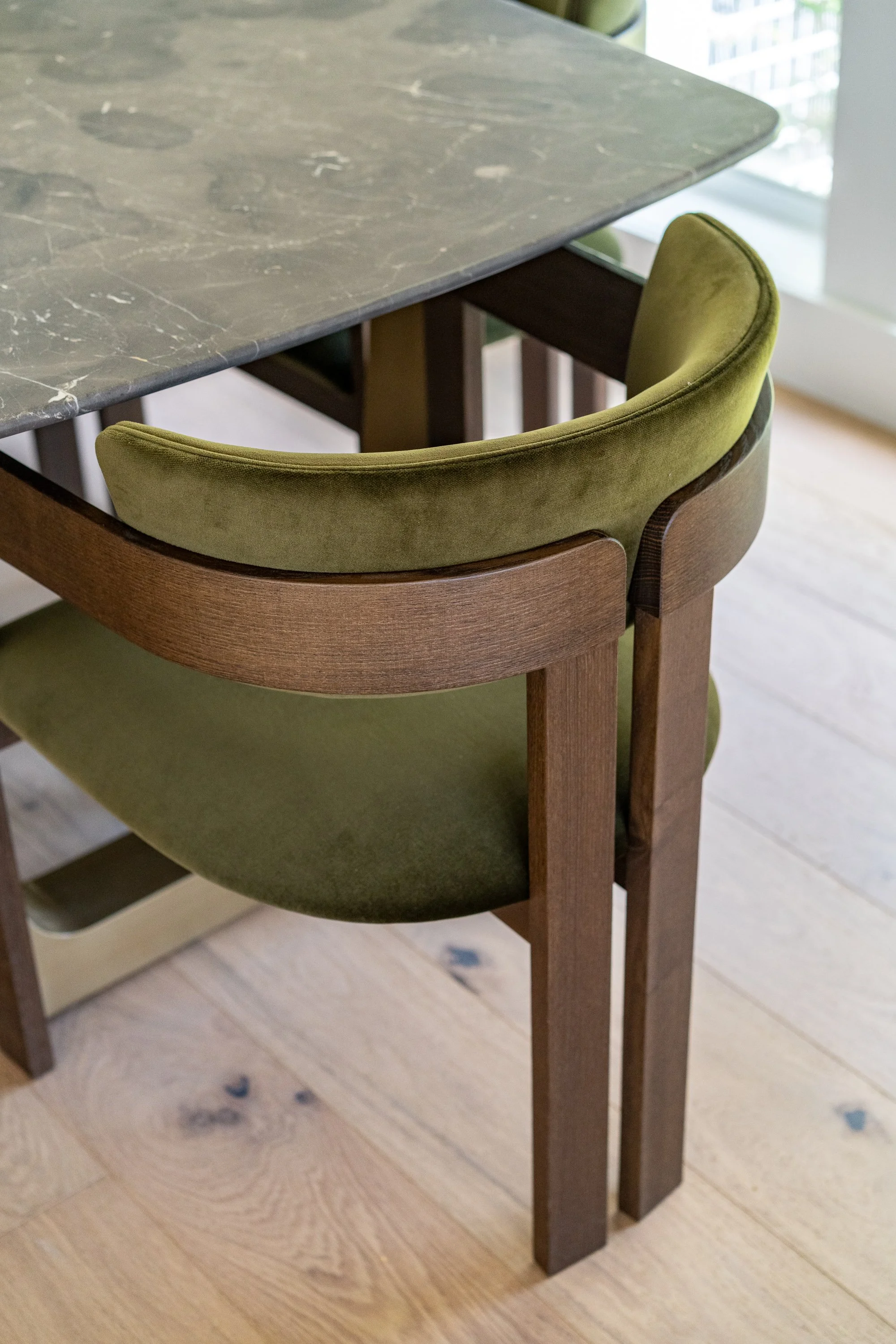 Close-up of a wooden dining chair with green upholstered seat and backrest, positioned next to a marble slab table, near a window.