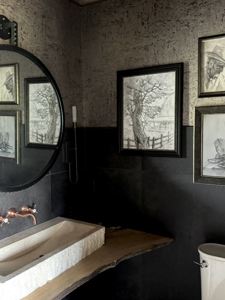A rustic bathroom with a stone sink, copper faucet, circular mirror, black and gray framed artwork of trees and boots, and a portion of a white toilet