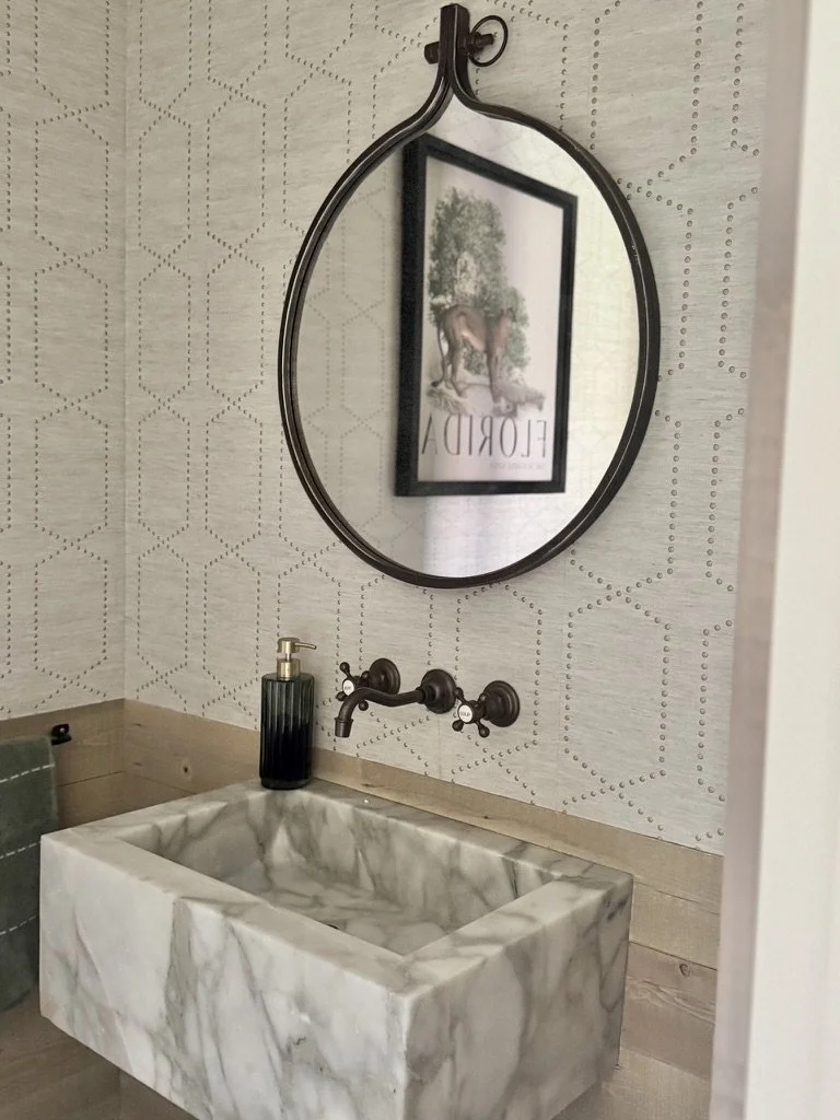 Bathroom with a marble sink, wall-mounted faucet, a bottle of soap, a hexagon patterned wallpaper, and a framed mirror reflecting artwork with the word 'Florida' and a horse.