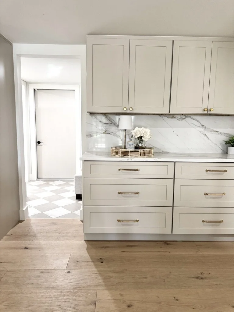 A section of a kitchen with white cabinets, a marble backsplash, and a wooden countertop. There is a decorative tray with a flower arrangement and a small lamp. The floor is wooden, and a doorway with a checkered tile floor is visible in the backgrou