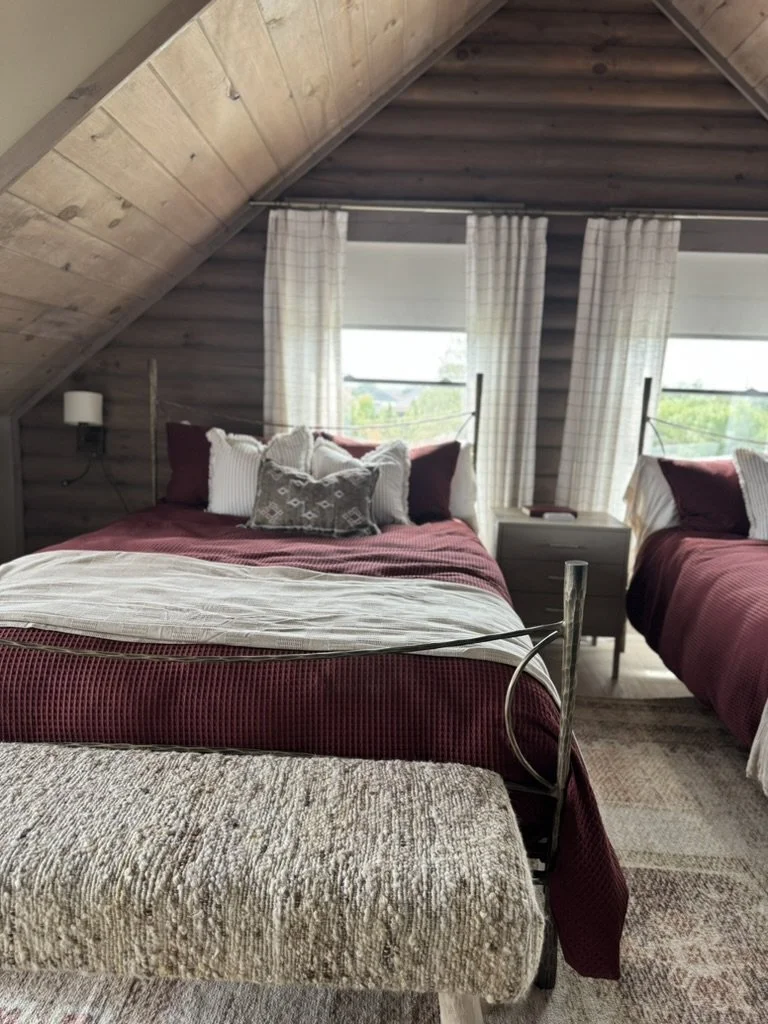 Two beds with burgundy and white bedding in an attic bedroom with wooden walls and sloped ceiling, beige curtains, and a window showing greenery outside.