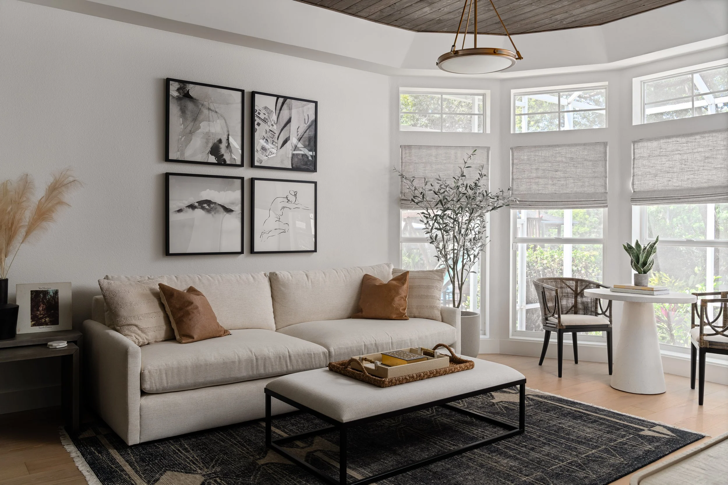 Living room with a beige sofa, brown pillows, black and white framed artwork on the wall, a black side table, a large potted plant, a round white table with a plant and books, two chairs, and a window with blinds.