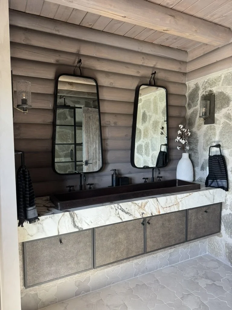 A rustic bathroom with a double sink vanity featuring a marble countertop and two large, black-framed mirrors. There are black faucets, a white vase with flowers, black towels, and wall-mounted light fixtures on a stone wall.