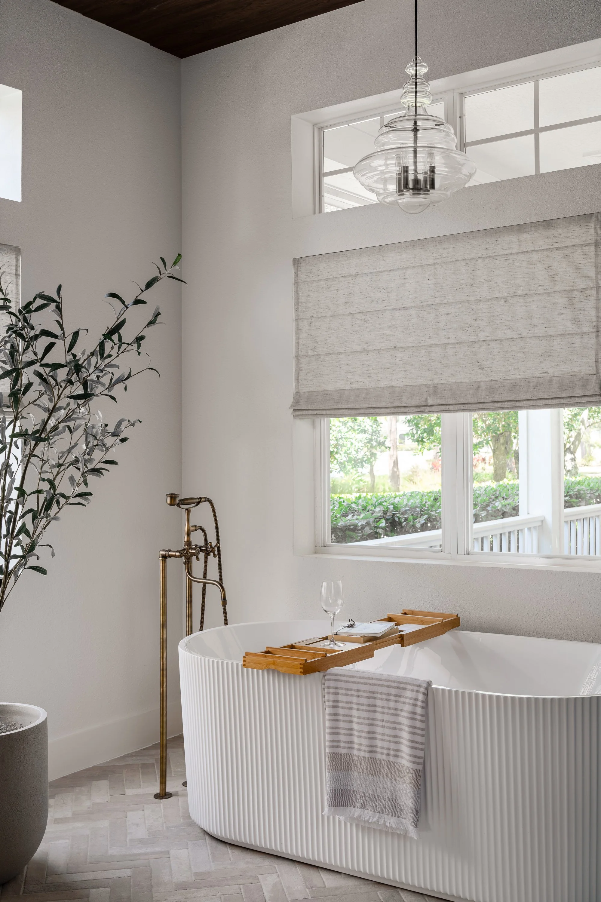 A modern bathroom with a white freestanding bathtub in front of a large window, a wooden bath tray with a wine glass, a striped towel hanging on the bathtub, a potted plant, and a glass pendant light hanging from the ceiling.