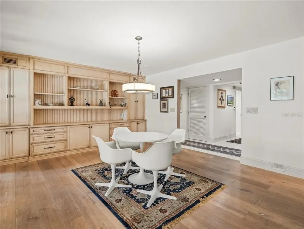 Dining room with a round white table, four white chairs, a wooden built-in wall cabinet, a hanging light fixture, and a patterned rug on wooden flooring. White walls decorated with framed pictures.