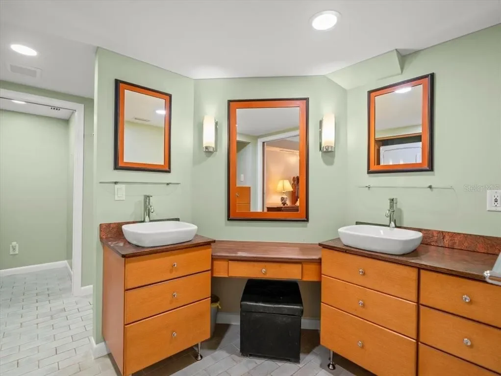 A bathroom with green walls, two wooden vanities with vessel sinks, and matching framed mirrors. There are wall-mounted light fixtures next to each mirror. The floor is tiled, and there's a doorway leading to another room with a lit lamp.