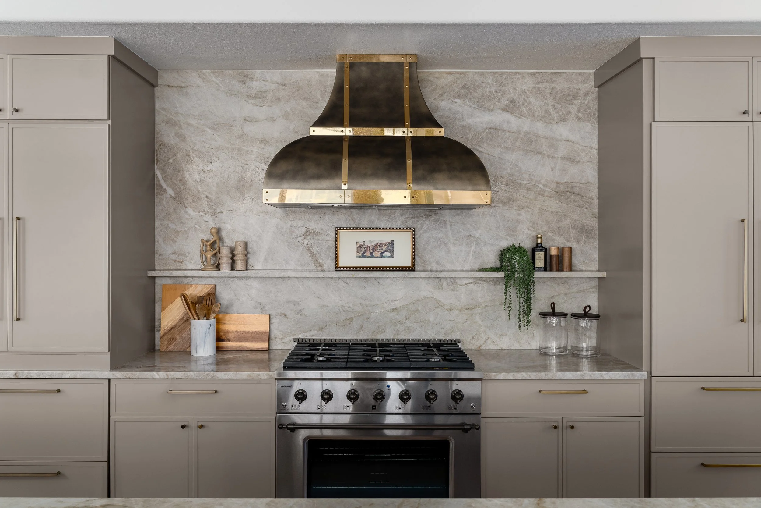 Modern kitchen with a stove, beige cabinets, marble backsplash, brass handles, decorative items on the shelf including small sculptures, a framed picture, potted plant, and storage jars, above a marble countertop and range hood.
