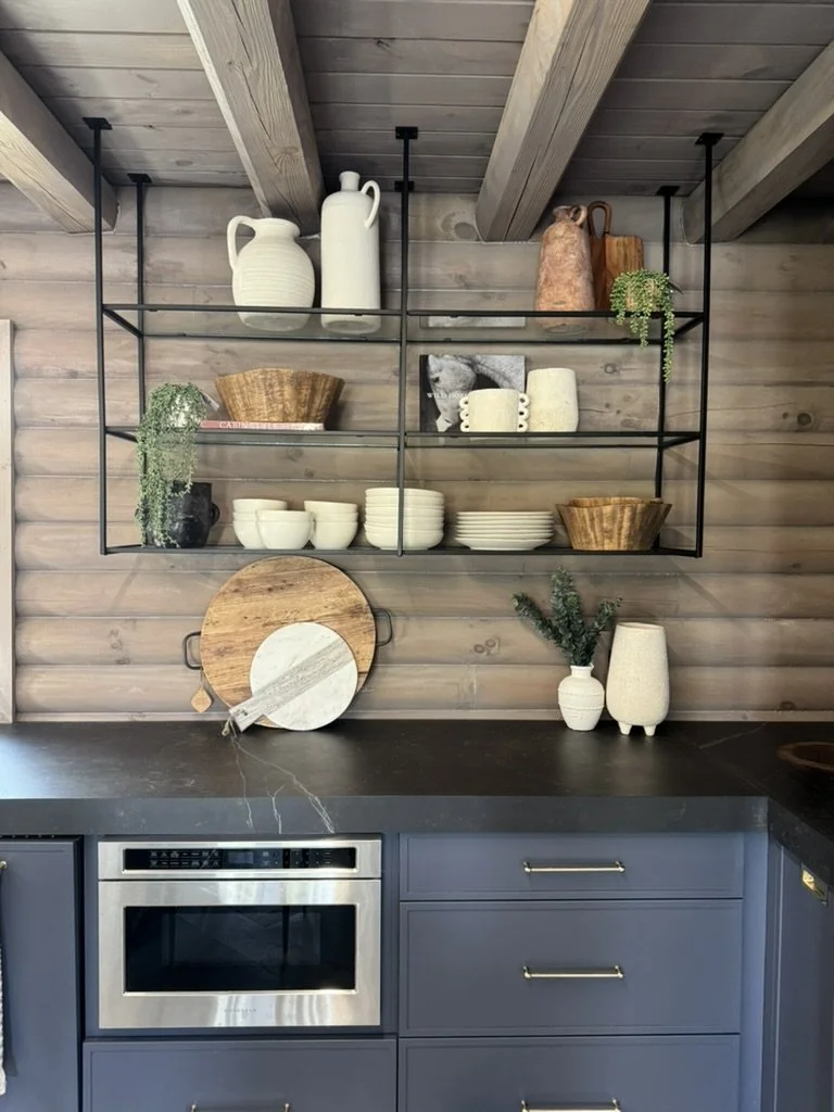 Open black metal shelving unit with white pitchers, brown and beige vases, green plants, white bowls, and wooden bowls, mounted on a wood-paneled wall above a black countertop with a blue cabinet and stainless steel oven.