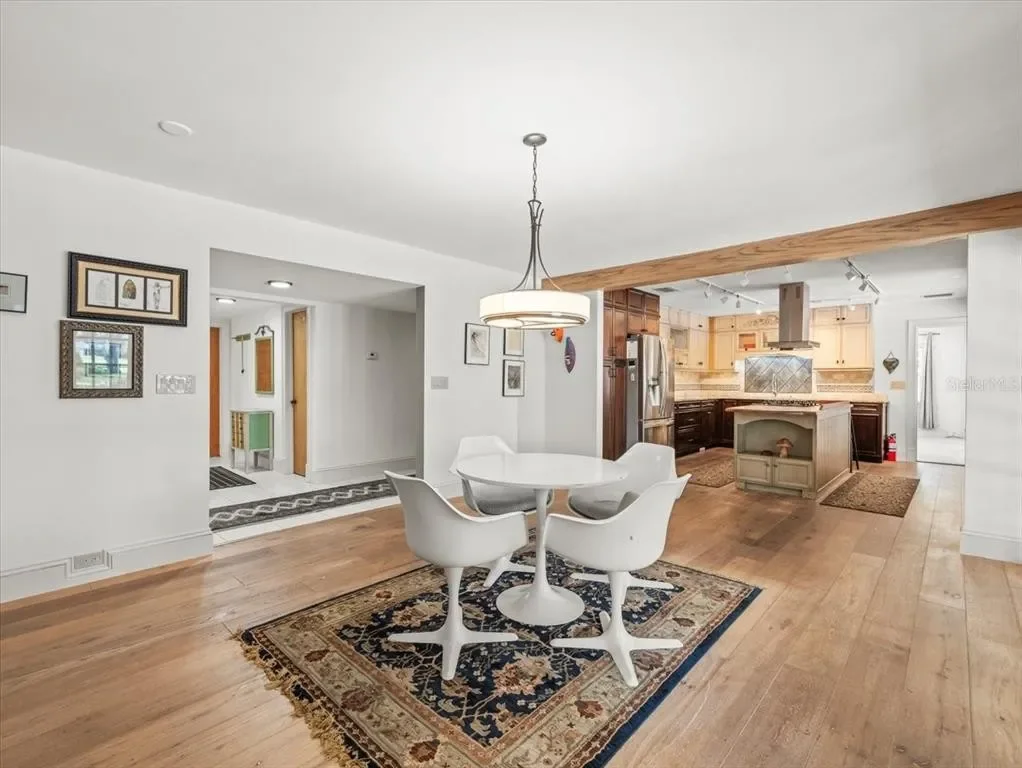 Open-concept dining area with a white round table and four white modern chairs on a decorative rug, adjacent to a kitchen with wooden and dark cabinetry and a kitchen island with a small display shelf, all in a bright, white-walled room with wooden f