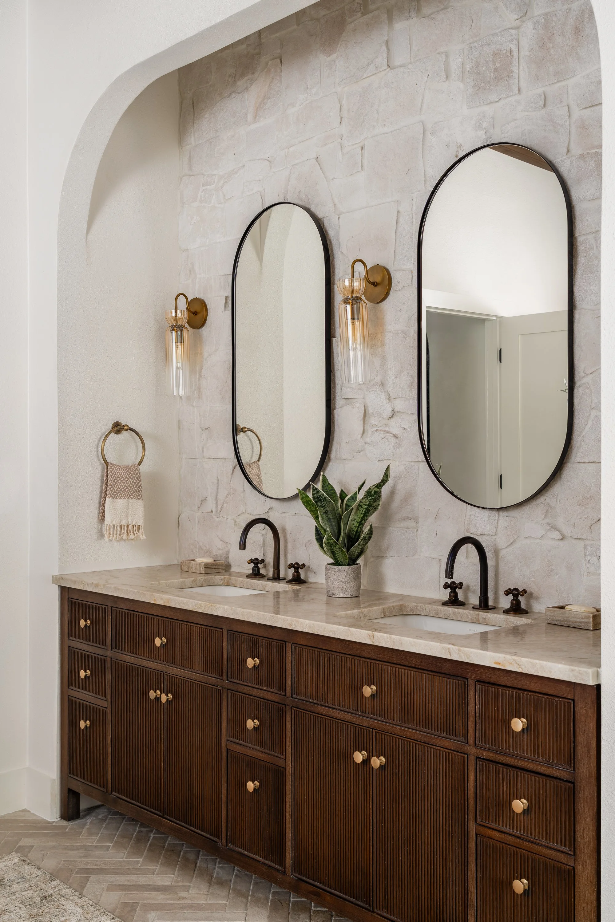 Bathroom vanity with two sinks, two oval mirrors, wall-mounted light fixtures, a potted plant, a towel, and a multi-drawer wooden cabinet.