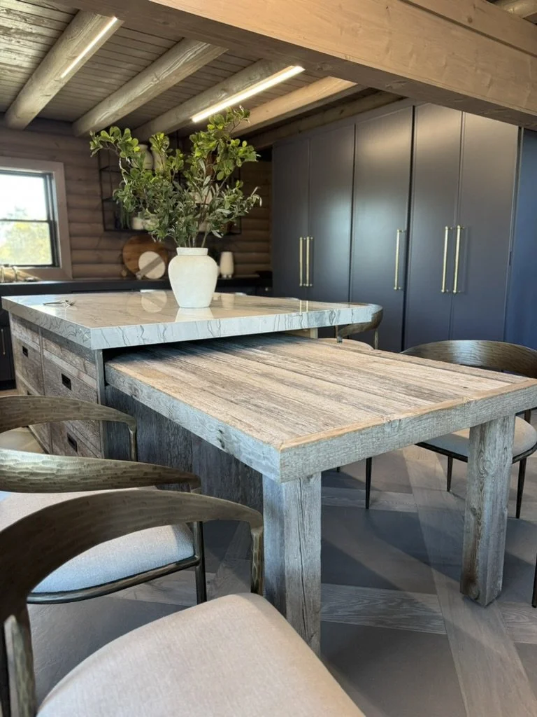 A cozy kitchen with a wooden dining table, a marble countertop, and a potted plant on the island. Blue cabinets with gold handles are visible in the background, along with a window and wooden ceiling beams.