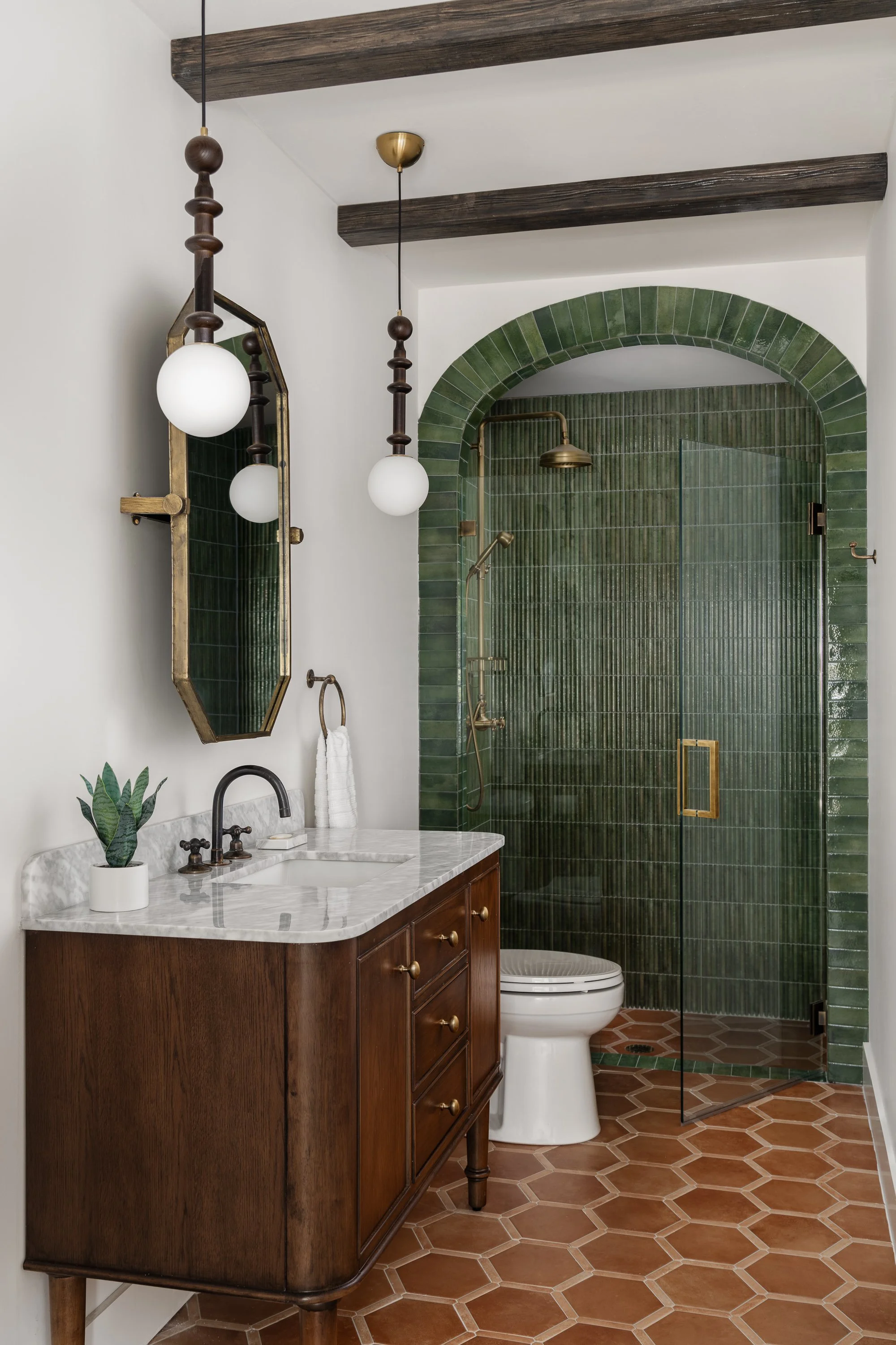 A bathroom with a wooden vanity, marble countertop, and a potted plant. Behind the toilet is a green tiled walk-in shower with a glass door and brass fixtures. There are two pendant lights hanging from the ceiling.