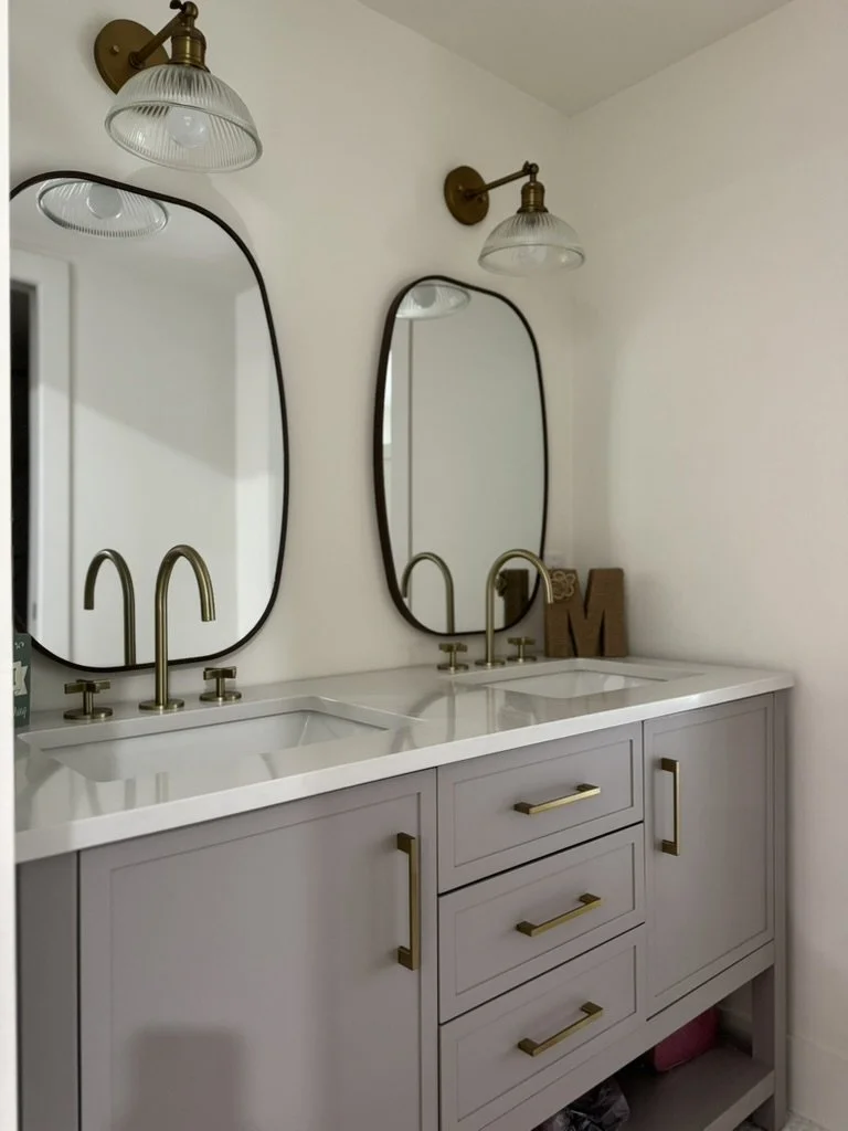 A bathroom vanity with a white countertop, two sinks, and three drawers, topped with two oval mirrors and two brass wall-mounted light fixtures.