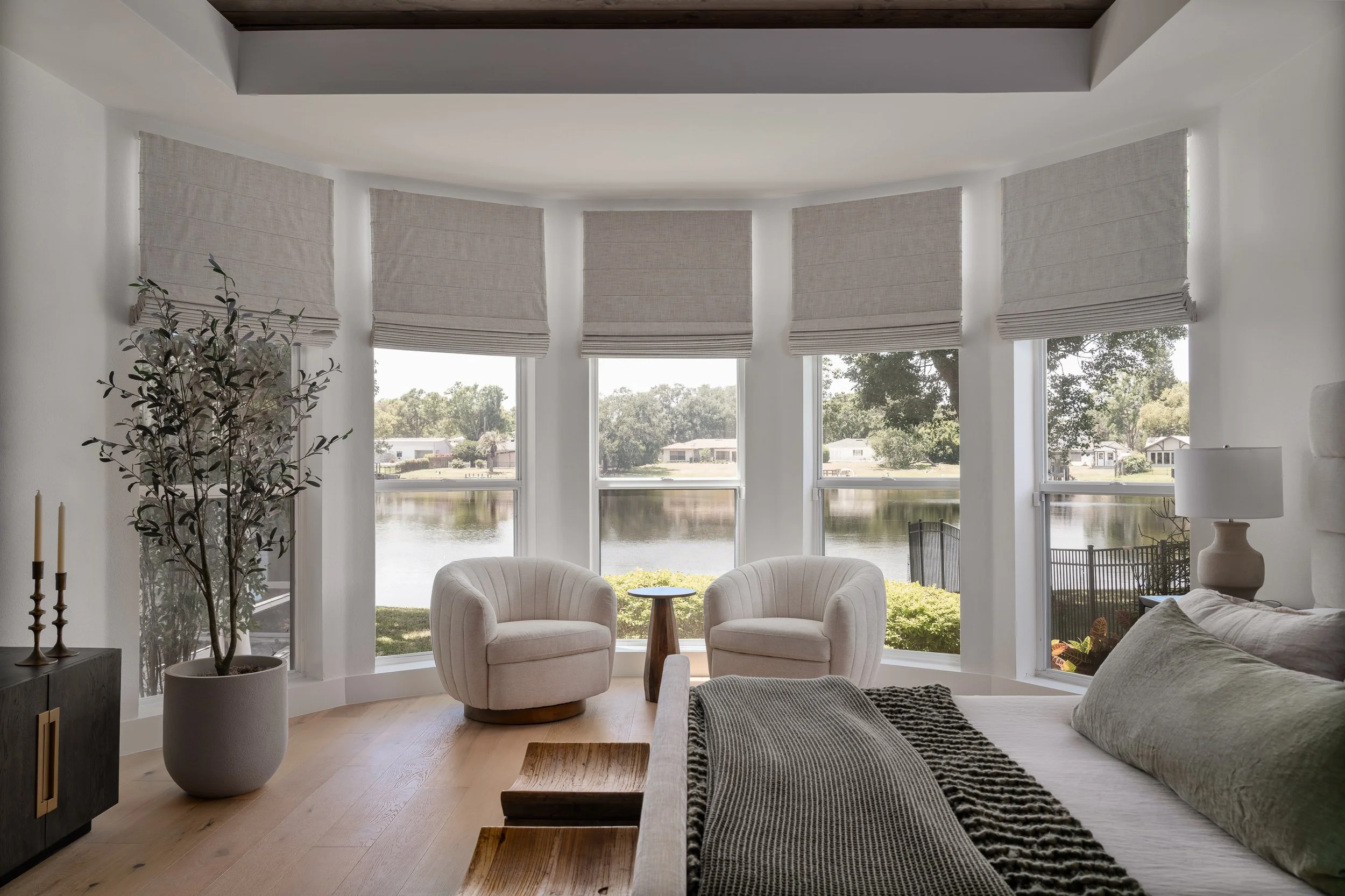 Living room with large bay windows overlooking a lake, two beige armchairs, a small wooden side table, a bed with green pillows and a dark bedspread, a white table lamp, a potted plant, and a black sideboard.