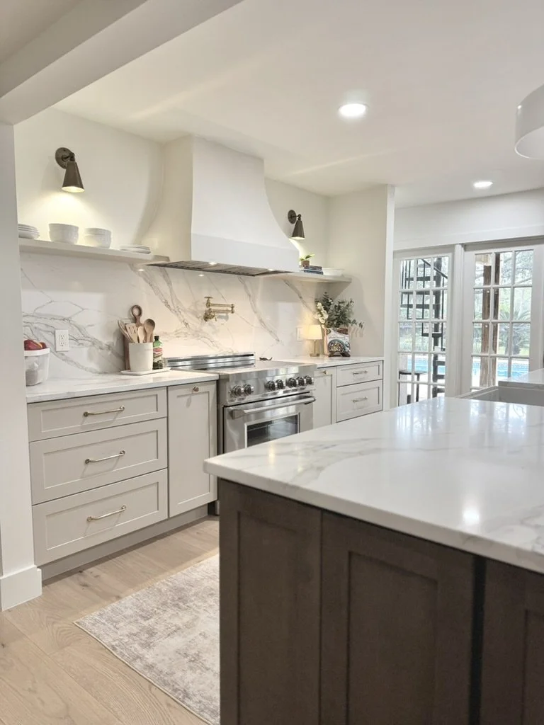 Modern kitchen with white cabinetry, marble countertops, stainless steel stove, and open shelves displaying dishes and decor, near sliding glass doors.