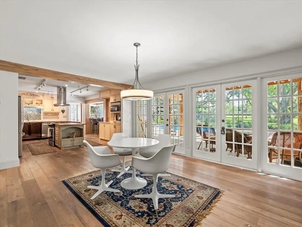 Open-concept dining area with a round white table, four white chairs, a patterned rug, and large glass doors leading to a porch with outdoor seating and lush greenery.