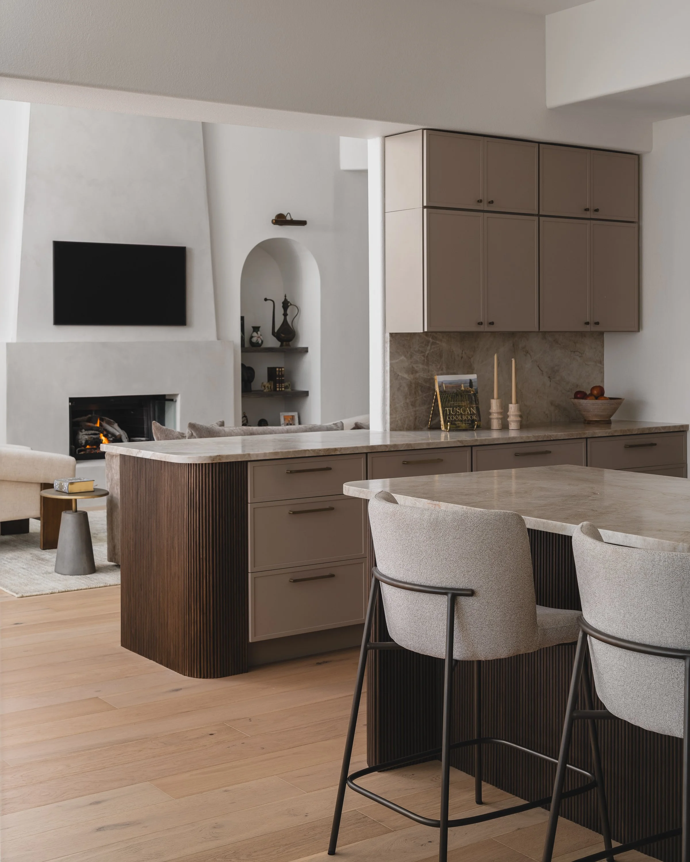 Modern kitchen with beige cabinetry, marble countertops, and a dining island with chairs. In the background, a living area with a mounted TV and a fireplace.