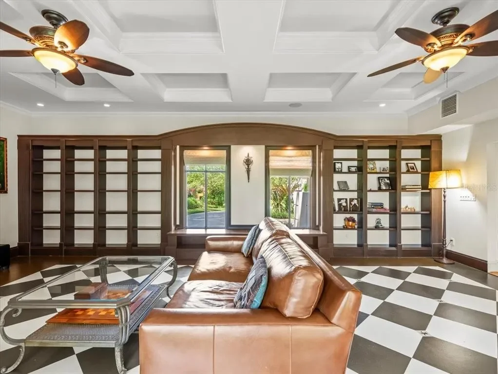 Living room with a leather sofa, a glass coffee table, window with a garden view, bookshelf, ceiling fans, and a floor lamp.