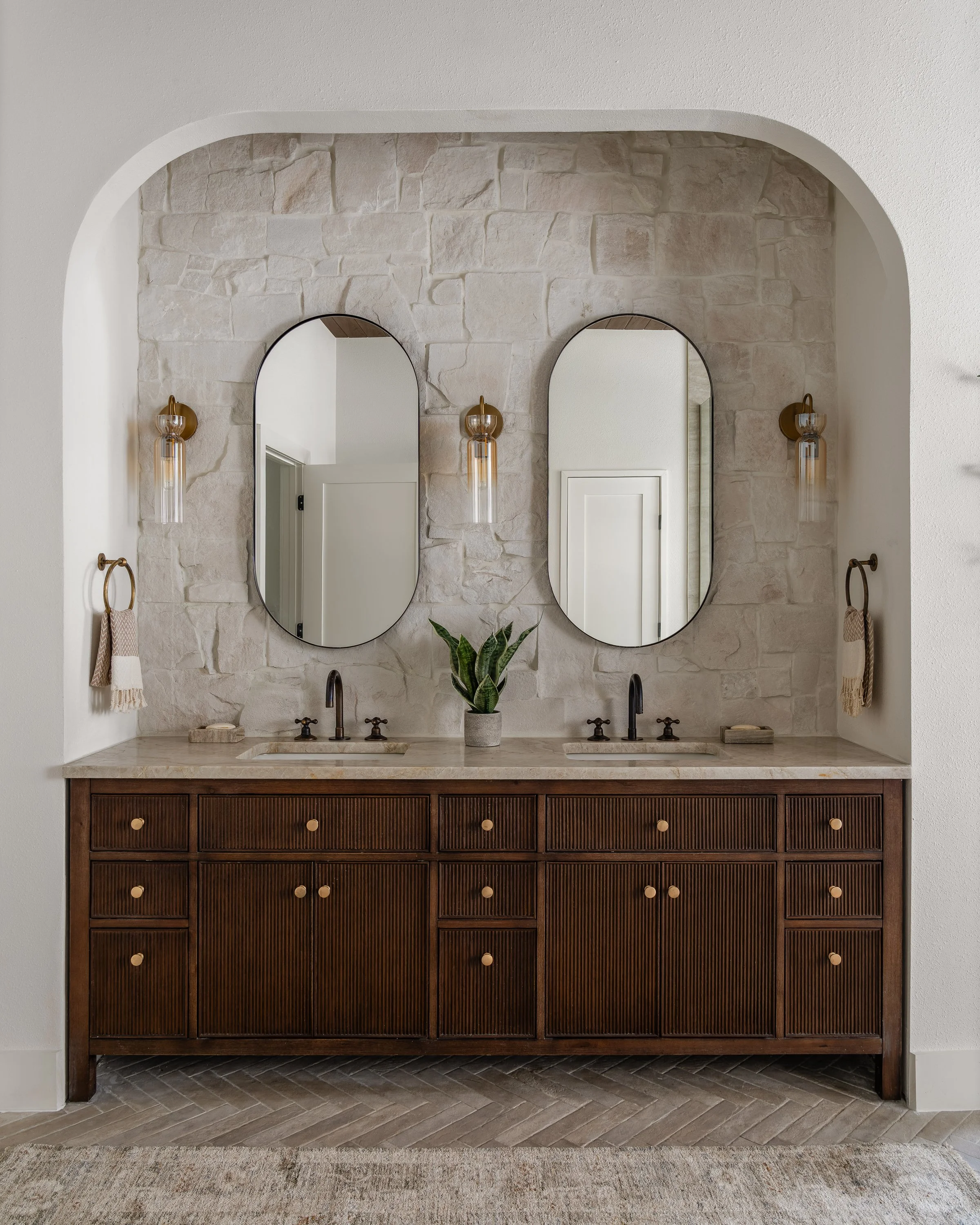 A modern bathroom vanity with a wooden cabinet, two sinks, and two oval mirrors mounted on a textured stone wall. Two wall-mounted light fixtures and towel rings with beige towels are on either side of the mirrors, with a potted plant in the center.