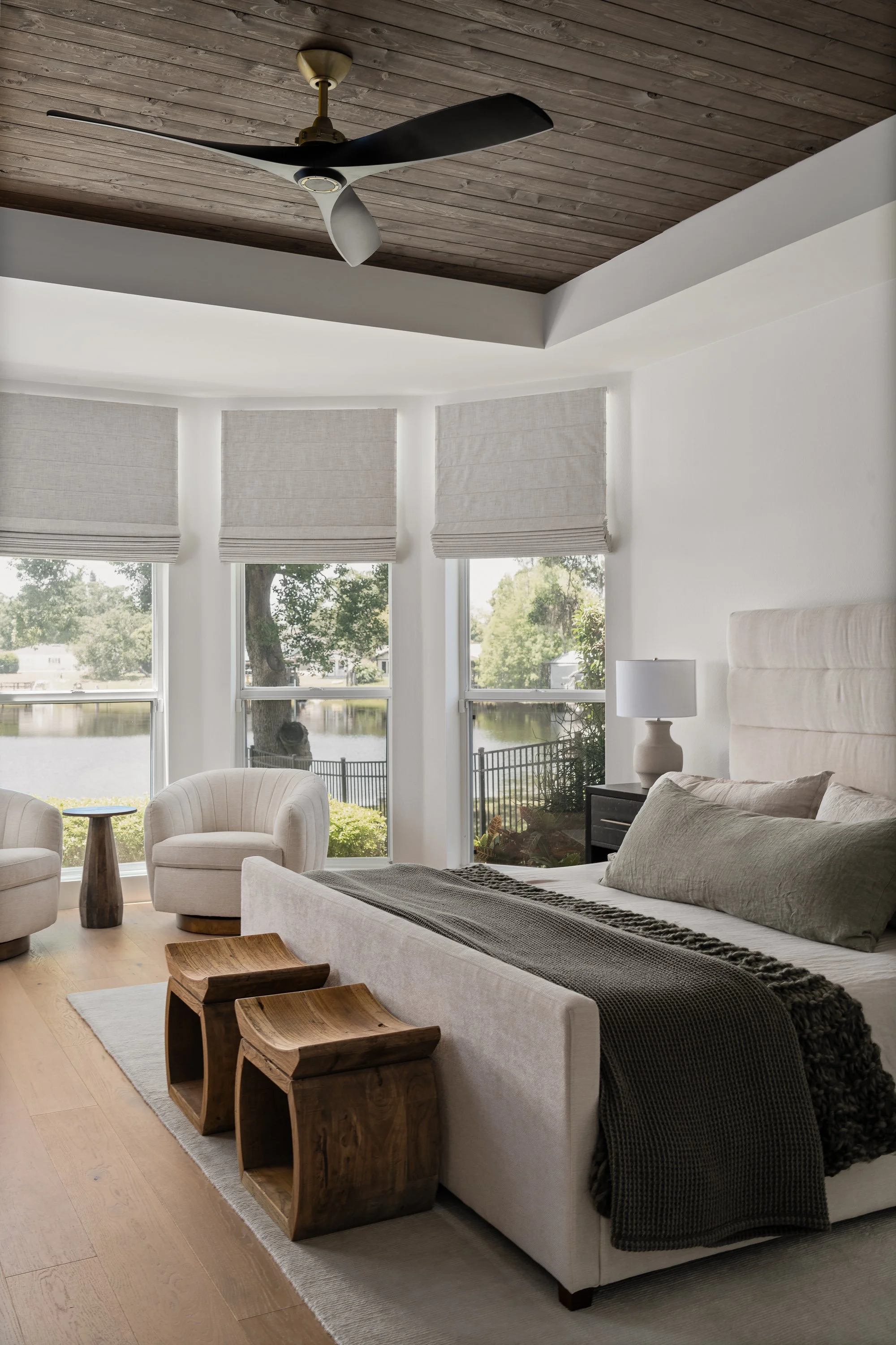 A bedroom with large windows overlooking a lake and trees, featuring a beige upholstered bed with pillows and a dark throw blanket, white walls, wood floors, two wooden stools at the foot of the bed, a nightstand with a lamp, and two cream-colored ch