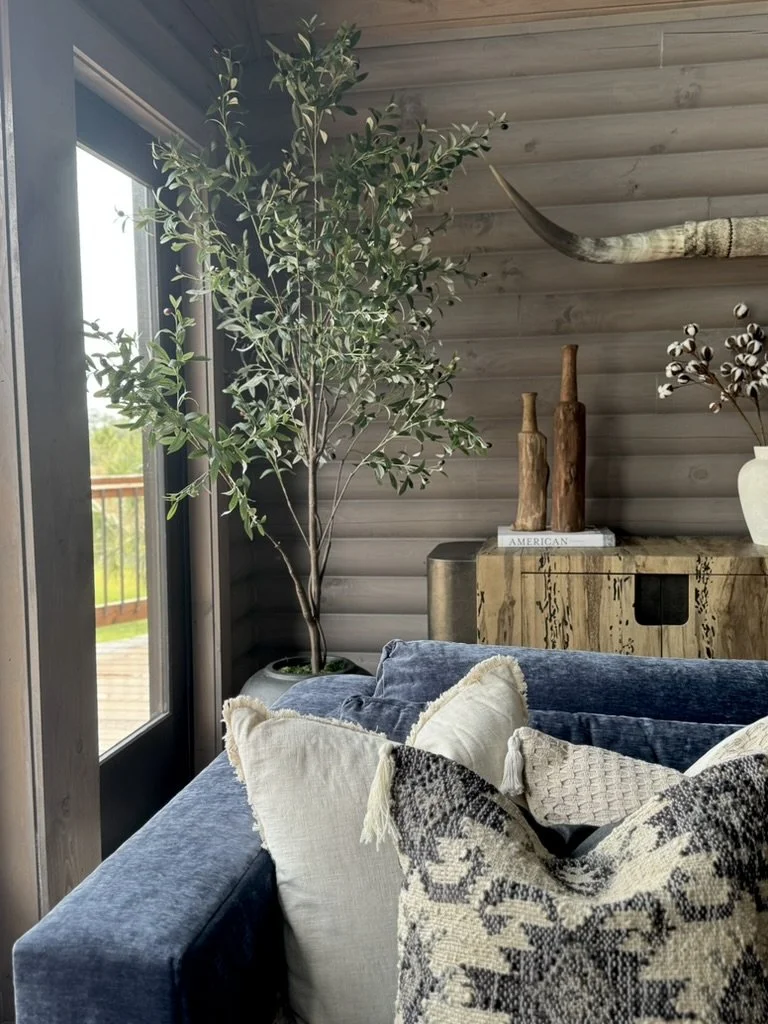 Living room corner with a large potted plant next to a window, a blue velvet sofa with several decorative pillows, and a rustic wooden sideboard decorated with vases and a book titled 'American'.