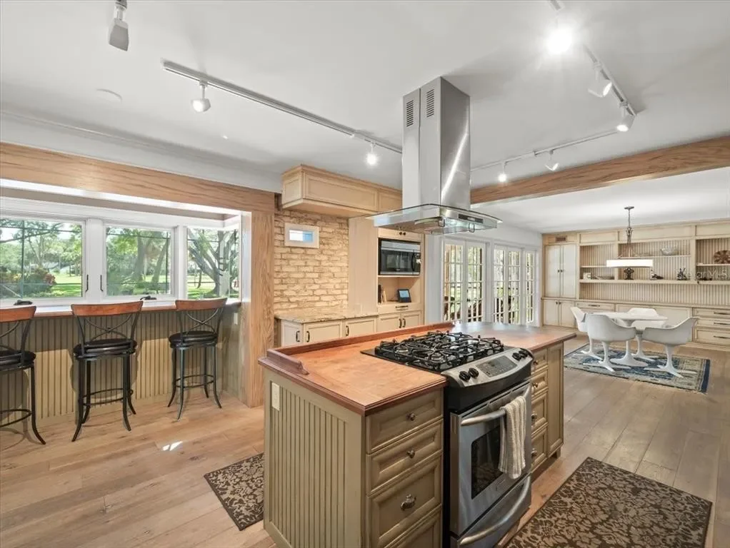 Open concept kitchen and dining area with wooden accents, large windows, and a kitchen island with a stove, stainless steel range hood, and seating area.