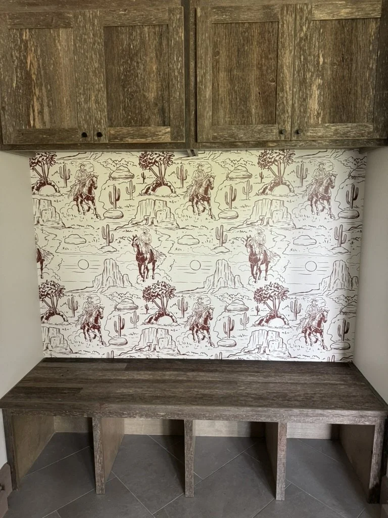 A rustic entryway with a wooden bench and rustic cabinets above. The wall has a country-themed wallpaper featuring scenes of cowboys riding horses, cacti, and desert landscapes with mountains and setting suns.