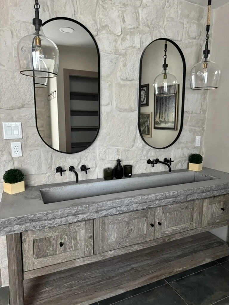 Modern bathroom vanity with a rustic wood finish, a built-in stone sink, two black wall-mounted faucets, and two oval mirrors. Hanging pendant lights and small potted plants are also visible.