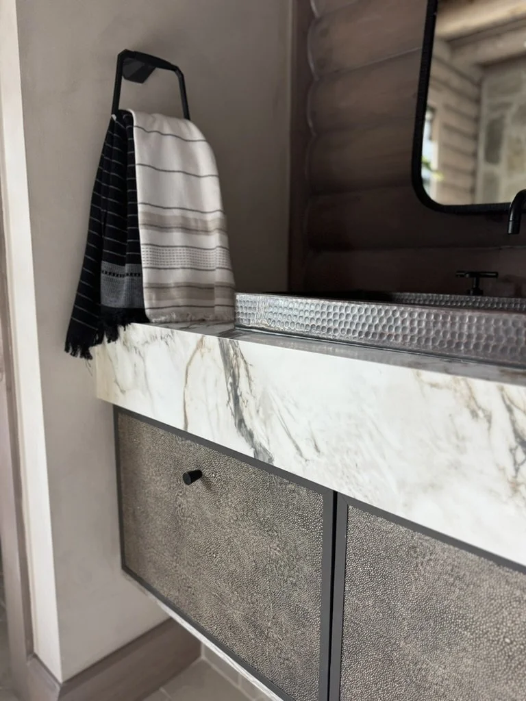 A bathroom vanity with a white marble countertop and textured cabinet doors. A black and white striped towel hangs on a black hook beside a mirror.