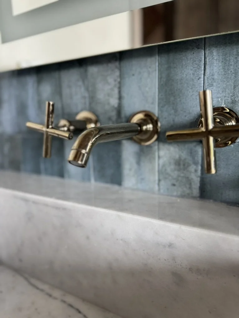 Close-up of two brass faucet handles and a brass spout above a white marble sink with a blue tiled backsplash.