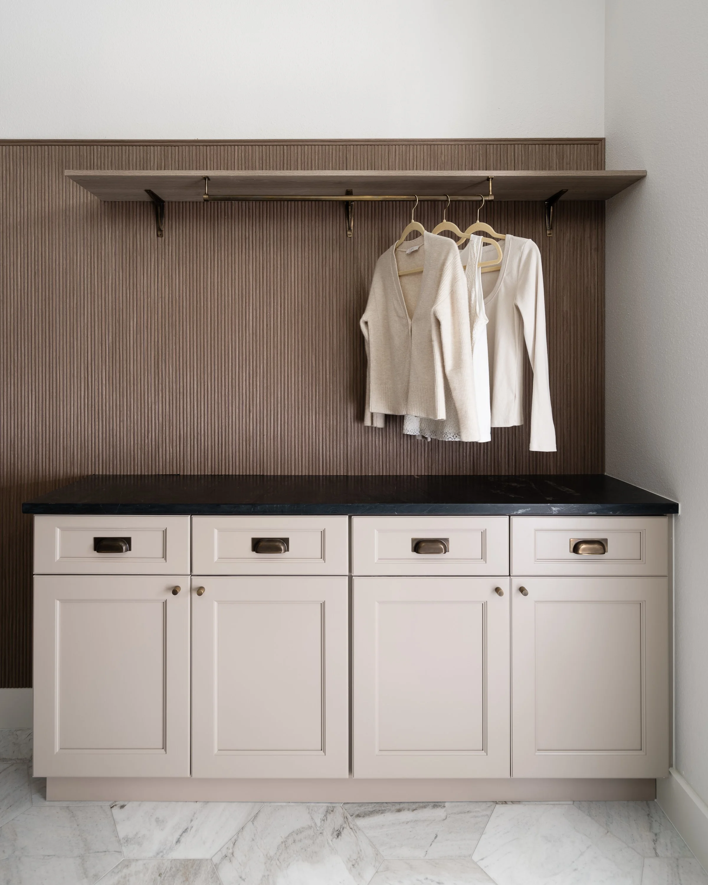 White cabinet with black countertop against brown, grooved wall, with three beige clothing hangers holding a beige vest, white blazer, and a white long-sleeve top, in a minimalist interior.