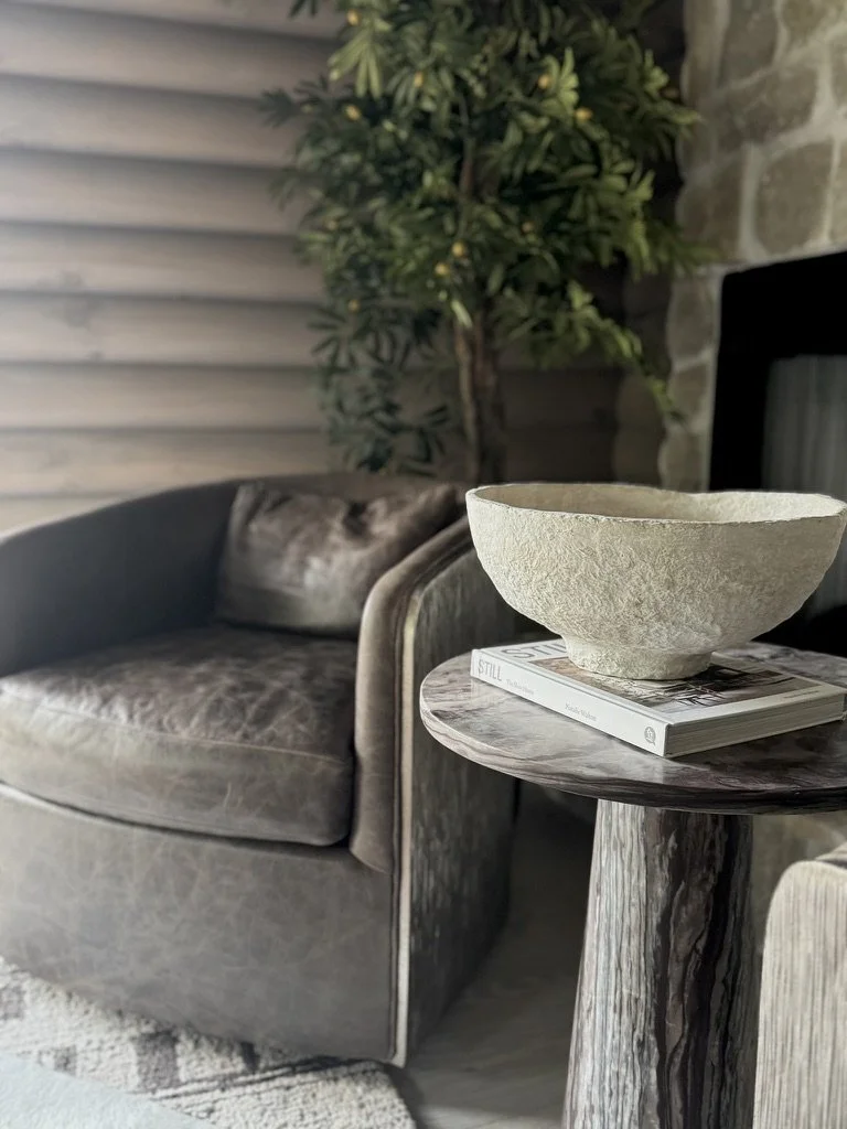 A cozy living room corner features a rust-colored velvet armchair, a small round side table with a marble pattern, a large stone bowl and a magazine, with a green potted tree and a stone wall in the background.