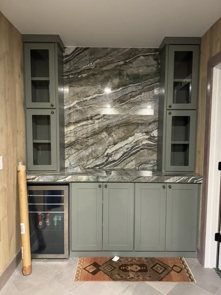 Home bar area with green cabinets, marble countertop and a marble backsplash, small wine fridge, and a colorful rug on the tiled floor.