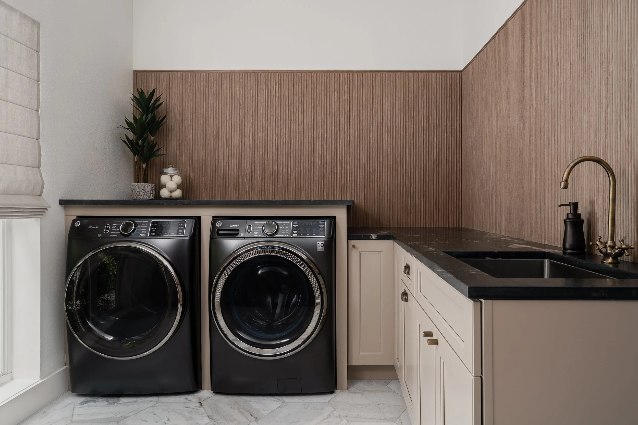 Laundry room with front-loading washer and dryer, black countertop, beige cabinets, and a black sink with gold faucet.