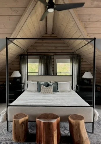 A cozy bedroom with a pitched wood ceiling, a four-poster bed with white bedding and pillows, dark nightstands with lamps, and three wooden stools in front of the bed.